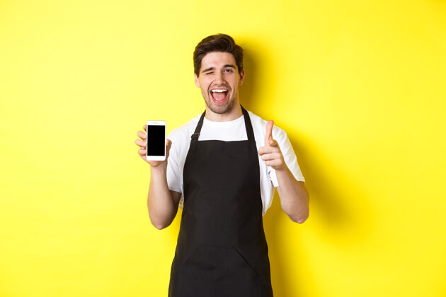 自信快乐的服务员展示手机屏幕 竖起大拇指 推荐咖啡馆餐厅应用程序 站在黄色背景上男士男士成人