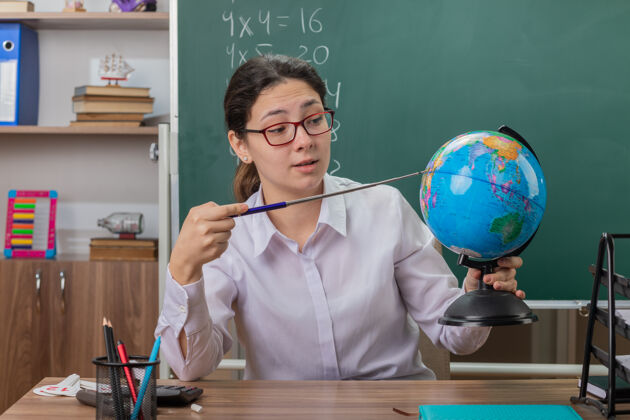 老师年轻的女老师戴着眼镜拿着地球仪和指针在教室里讲课 自信地坐在黑板前的课桌旁桌子前面拿着