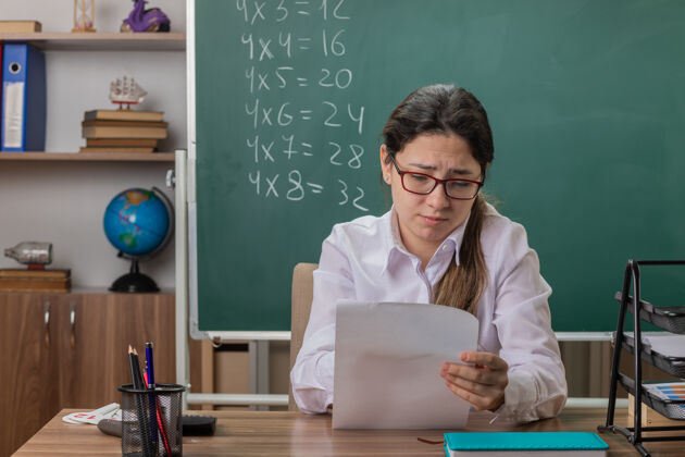 教室戴着眼镜的年轻女教师坐在教室的黑板前 一副茫然而不高兴的样子坐着戴着眼镜