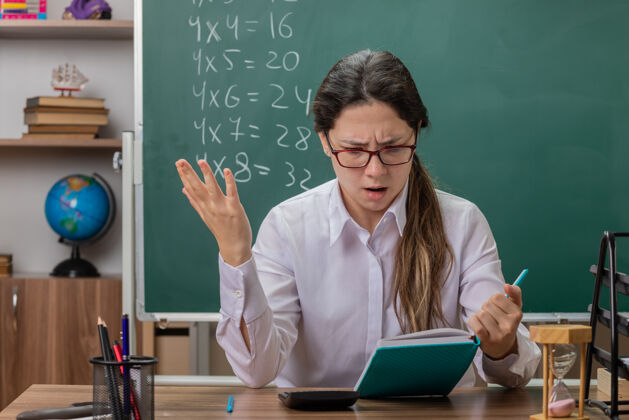 前面年轻的女老师戴着眼镜拿着笔记本教室黑板书桌