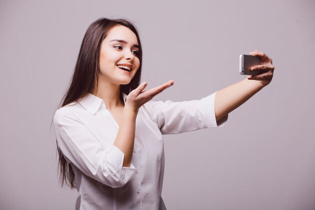 相机在白色背景下 一个快乐的调情的年轻女子在智能手机前拍下了自己吹吻的照片摄影师手机爱