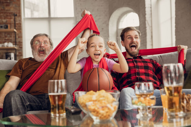 篮球胜利兴奋 快乐的家庭看篮球比赛 冠军在家里的沙发上球迷为最喜爱的国家队情绪欢呼女儿 爸爸和爷爷体育 电视 玩得开心微笑乐趣观看