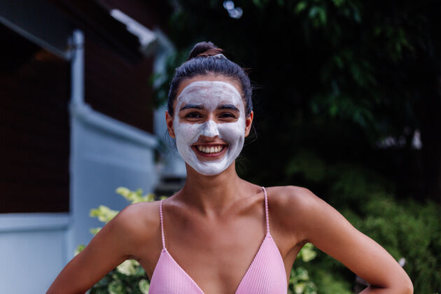护理晒黑皮肤的户外写真美丽的高加索女子穿着比基尼在温泉浴场与白色脱皮面具在脸上面部放松触摸