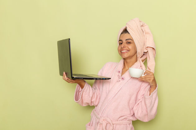 咖啡前视图穿着粉色浴袍的年轻女性使用笔记本电脑 在浅绿色表面上喝咖啡口罩饮料年轻女性
