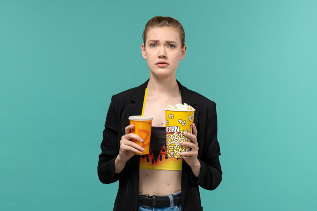 抱着正面图：年轻女性手持爆米花饮料 在浅蓝色的表面上看电影前面漂亮电影