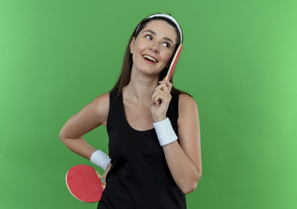 球拍戴着头巾拿着球拍打乒乓球的年轻健身女子站在蓝色的墙上 面带微笑地看着一边健身女人乒乓