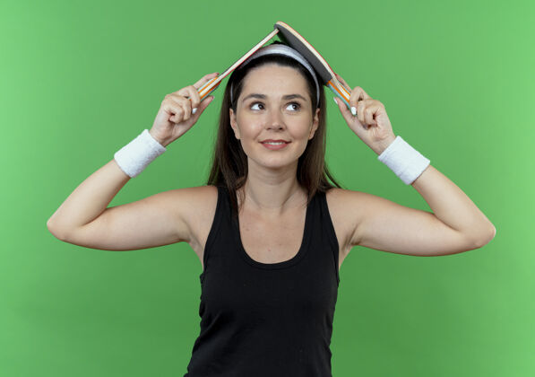 女子戴着头巾 头上举着两个乒乓球拍 站在绿色墙壁上微笑的年轻健身女士头部抱着头带