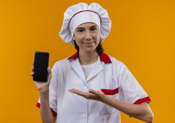 请身着厨师制服的年轻高加索厨师女孩高兴地拿着电话 指着橙色空间里的电话 还有复印空间点厨师制服