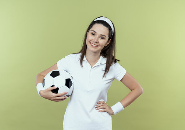 臀部戴着头巾的年轻健身女 手举足球 臀部微笑着 快乐而积极地站在轻质的墙上微笑女子足球