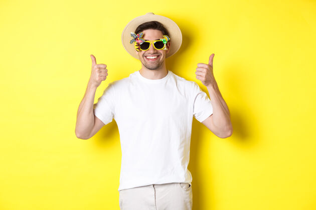 帅气旅游和生活方式的概念微笑的游客竖起大拇指 享受旅行和推荐 戴着夏季帽子和太阳镜 黄色背景的形象自信男人夏天