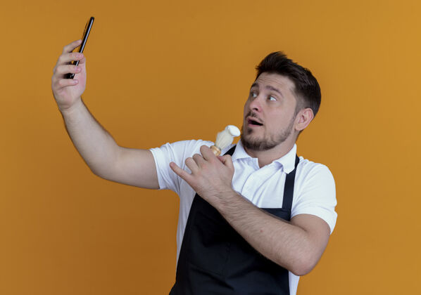 泡沫围裙上的理发师拿着剃须刷在脸上抹剃须泡沫 站在橙色的墙上用智能手机自拍用拿着理发师