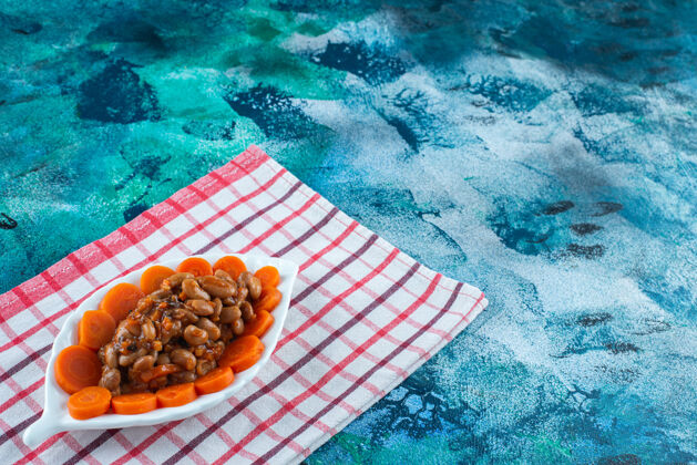 盘子把切成片的胡萝卜和豆子放在茶巾上的盘子里 放在蓝色的背景上切片豆类可口