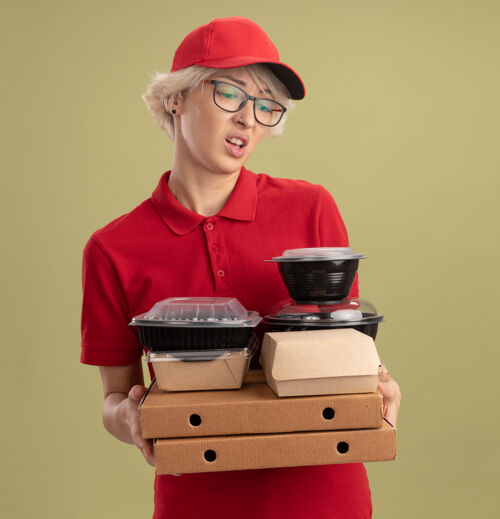 拿着年轻的送货员身穿红色制服 戴着帽子 戴着眼镜 手里拿着比萨饼盒和食品包 站在绿色的墙上 表情悲伤地看着它们披萨食物悲伤