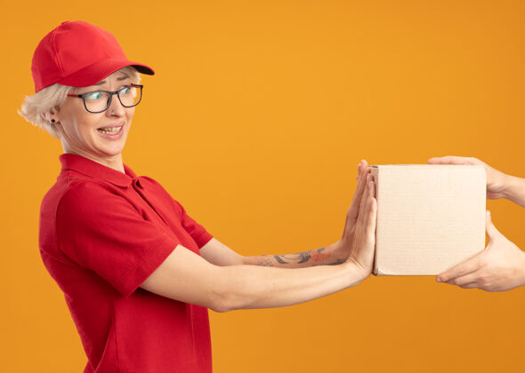 拿着身穿红色制服 戴着帽子的年轻送货员站在橙色的墙上拒绝拿箱子拒绝包装制服