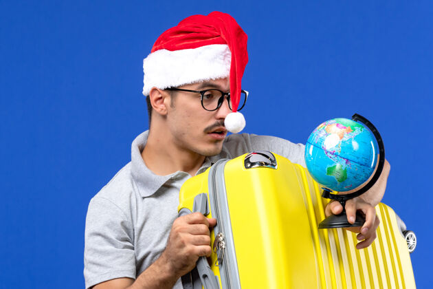 帽子前近景年轻男子手持地球仪和黄包在蓝墙飞机上度假旅行前面年轻男性人
