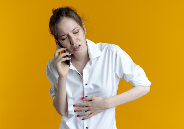 女孩年轻疼痛的金发俄罗斯女孩把手放在肚子上讲电话隔离在橙色空间与复制空间说话俄语放