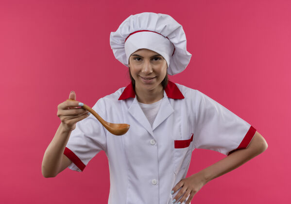 女孩穿着厨师制服的年轻快乐的白人厨师女孩拿着木勺子隔离在粉色空间和复印空间粉红年轻勺子