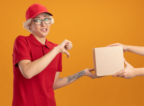 站着身穿红色制服 头戴帽子的年轻送货员站在橘色的墙上 一边接受礼盒包装 一边不高兴地竖起大拇指显示交货而