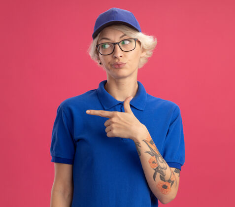 年轻不满意的年轻女送货员穿着蓝色制服 戴着帽子 困惑地向一旁望去 用食指指着站在粉红色墙上的一边帽子侧线女人