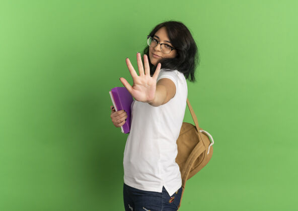 包年轻漂亮的白人女生戴着眼镜 背着书包 手拿着牌子 手拿着书 在绿色的空间里 有一个单独的复印空间请手背部