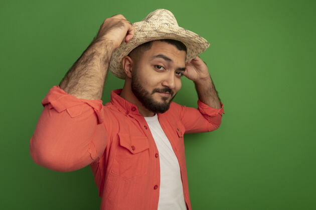 自信年轻的留着胡子的男人穿着橙色衬衫 戴着夏天的帽子 站在绿色的墙上 微笑着自信地修理他的帽子微笑站立夏天