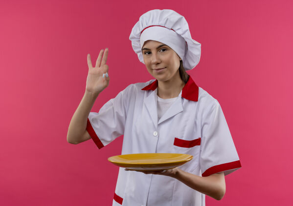 请年轻高兴的白人厨师女孩 身着厨师制服 拿着盘子 手势ok 手势孤立在粉色空间和复印空间拿着粉色空间