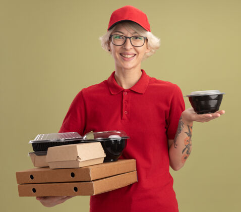 送货身穿红色制服 戴着眼镜 拿着比萨饼盒和食品包的年轻送货员站在绿色的墙上 面带笑容制服拿着年轻人