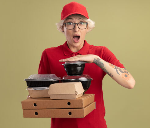 惊喜年轻的送货员身穿红色制服 戴着眼镜 手里拿着披萨盒和食品包 站在绿色的墙上 又高兴又惊讶穿上眼镜拿着
