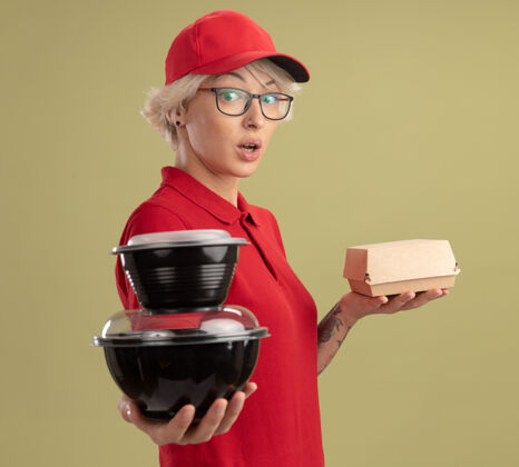 女人年轻的送货员身穿红色制服 戴着帽子 戴着眼镜 手里拿着食品包 站在绿色的墙上 看上去既惊讶又惊奇拿着年轻人惊喜