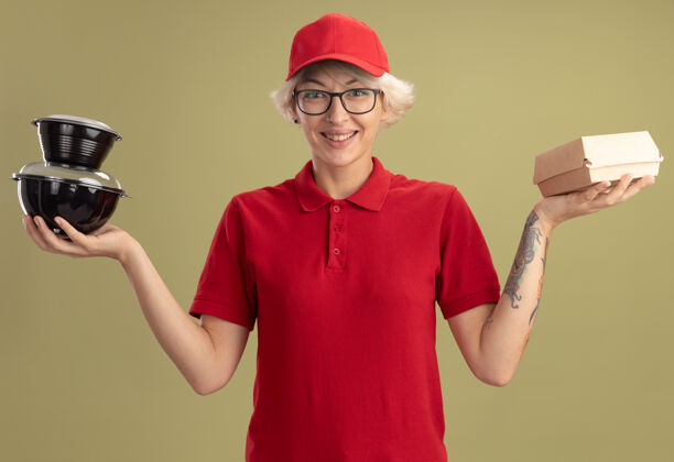 眼镜年轻的送货员身穿红色制服 戴着眼镜 手里拿着食品包 站在绿色的墙边开心地微笑着食物送货盒子