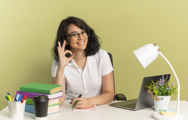 手势年轻的笑容可掬的高加索女学生戴着眼镜坐在书桌旁 拿着学习工具 手势ok 手语 拿着笔 在绿色空间和复印空间隔离开来绿色手可以