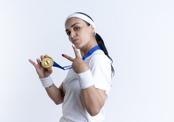 侧身年轻自信的白人运动女性戴着头带和腕带站在一边拿着金牌和手势胜利的手标志隔离在白色空间与复制空间穿运动手势