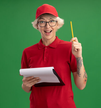 剪贴板身穿红色制服 戴着帽子 戴着一副眼镜 上面有空白页和铅笔的剪贴板的年轻女送货员站在绿色的墙壁上 高兴而兴奋地有了新的想法送货穿空白