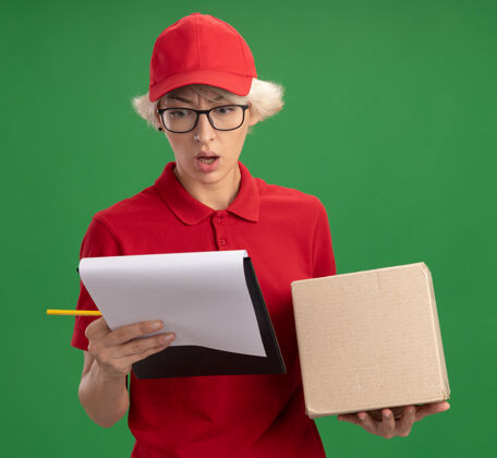 送货年轻的送货员身穿红色制服 戴着帽子 戴着眼镜 手里拿着纸板箱 手里拿着空白页的剪贴板 手里拿着铅笔 困惑地看着剪贴板站在绿色的墙上困惑铅笔拿着