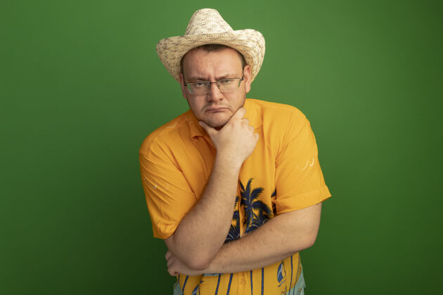 下巴戴着眼镜的男人 穿着橘色衬衫 戴着夏天的帽子 手放在下巴上 严肃的脸站在绿色的墙上思考戴手夏天