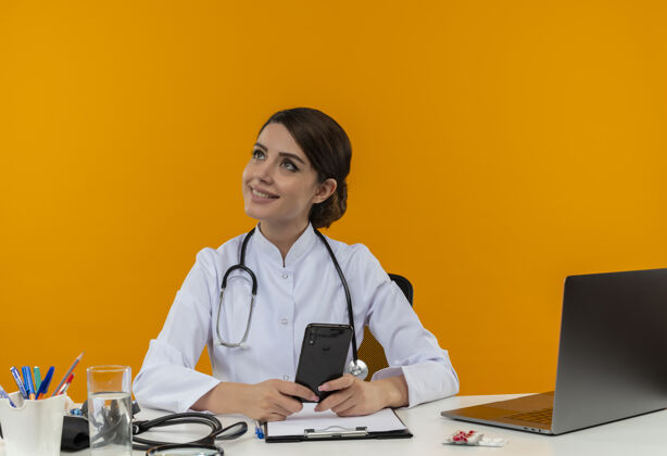 听诊器面带微笑的年轻女医生穿着医用长袍和听诊器坐在办公桌旁 手里拿着医疗工具和笔记本电脑 手里拿着手机 看着隔离在黄色墙上的一面女笔记本电脑坐着