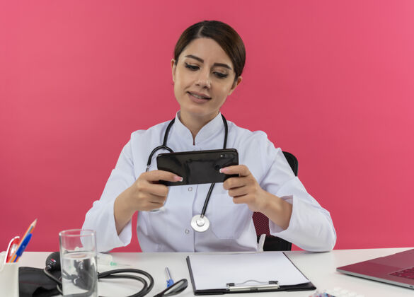 穿着年轻的女医生穿着医用长袍和听诊器坐在办公桌旁 手持医疗工具和笔记本电脑 用手机隔离在粉红色的墙上电话请办公桌