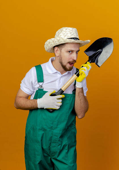 手套年轻英俊的斯拉夫园丁穿着制服 戴着帽子 戴着园艺手套 手里拿着铲子 看上去被隔离在橙色的墙上帽子斯拉夫穿