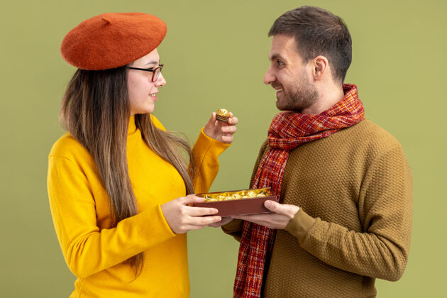 微笑年轻漂亮的夫妇幸福的男子提供巧克力糖果给他微笑可爱的女朋友在贝雷帽庆祝情人节站在绿色的墙壁男人女朋友糖果