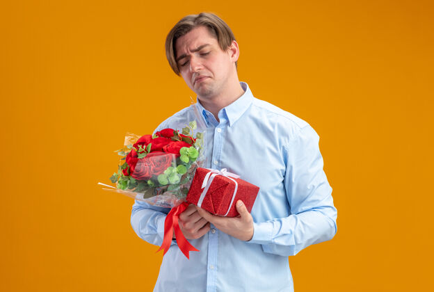 立场身穿蓝衣的悲伤男人手持一束红玫瑰 撅着嘴 带着失望的表情站在橘色的墙上 情人节的概念男人情人节概念