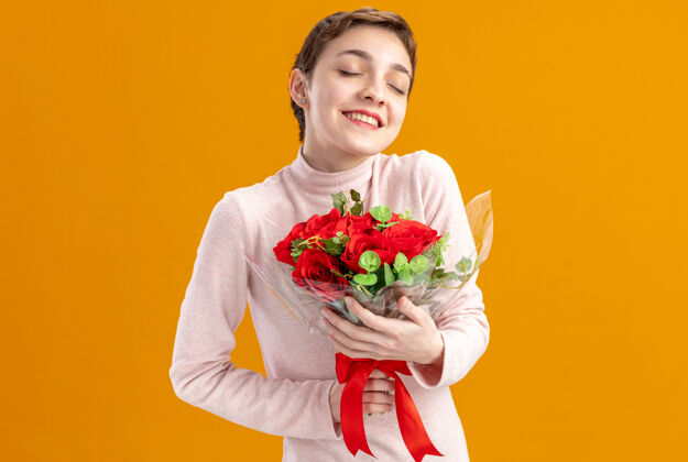 女人一个留着短发的年轻女子 手里捧着一束红玫瑰 快乐而乐观 闭着眼睛 微笑着站在橘色的墙上庆祝情人节微笑年轻立场