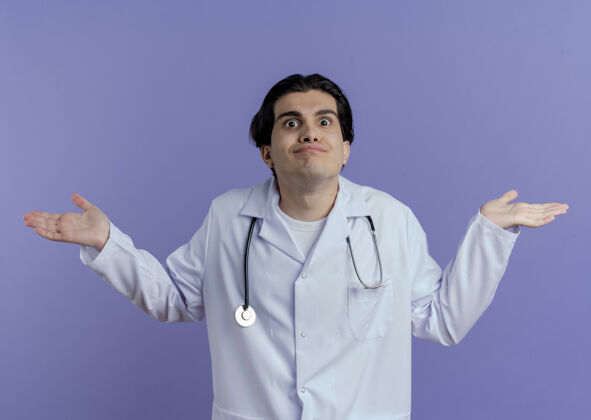 长袍困惑的年轻男医生穿着医用长袍和听诊器 双手空空的 孤立在紫色的墙上手显示医生