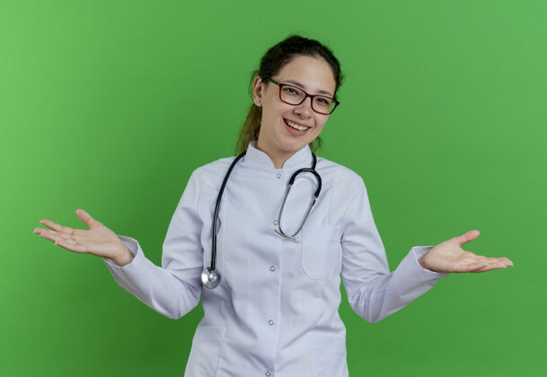 欢乐快乐的年轻女医生穿着医用长袍 戴着听诊器 戴着眼镜 双手空空的隔离在绿色的墙上女表演医生