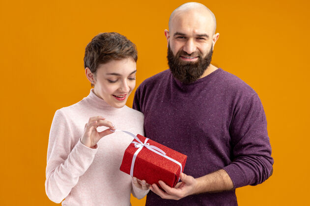 惊喜一对穿着休闲服的年轻夫妇微笑着 留着胡子的男人站在橙色的背景下 给他惊喜快乐的女友一份礼物衣服情侣女朋友