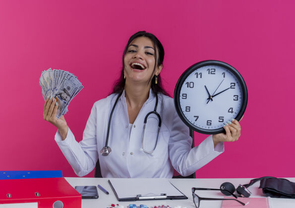 女性快乐的年轻女医生穿着医用长袍和听诊器坐在办公桌旁 手里拿着医疗工具 手里拿着钱 墙上的钟被隔离开来欢乐医疗长袍