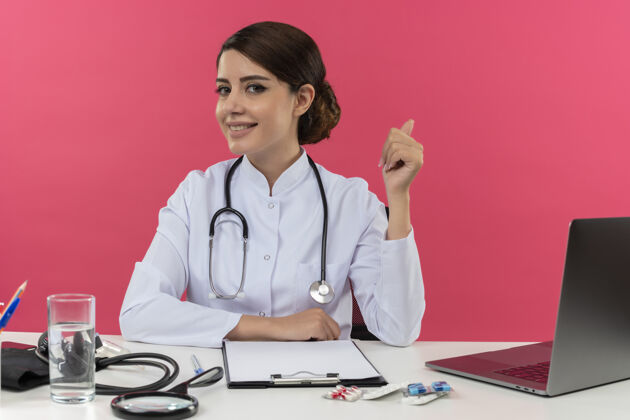 坐着微笑着的年轻女医生穿着医用长袍和听诊器坐在办公桌旁 手里拿着医疗工具和笔记本电脑 手放在办公桌上 把大拇指孤立地竖立在粉红色的墙上微笑医疗长袍