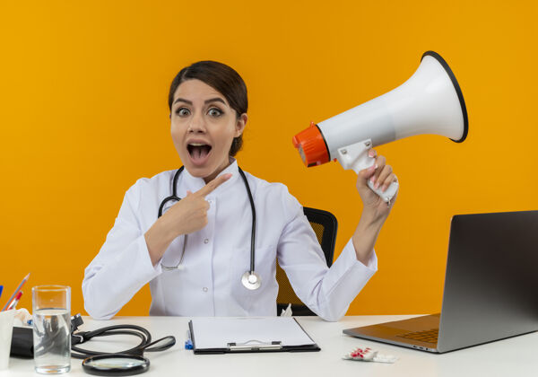 医疗令人印象深刻的年轻女医生穿着医用长袍和听诊器坐在办公桌旁 手持医疗工具和笔记本电脑 指着隔离在黄色墙上的扬声器指着女年轻