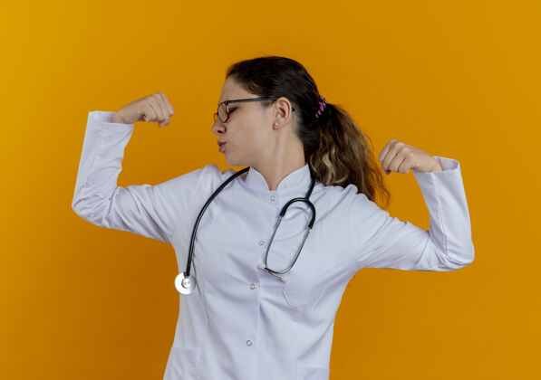 展示站在侧视图年轻的女医生穿着医用长袍和听诊器 戴着眼镜 显示出强烈的姿态隔离在橙色的墙上视野年轻手势