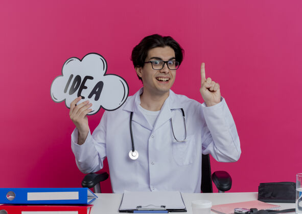 印象年轻的男医生穿着医用长袍 戴着听诊器 戴着眼镜 坐在办公桌旁 手里拿着医疗工具 手里拿着创意泡泡 举起手指孤立在粉红色的墙上坐着抱着医疗