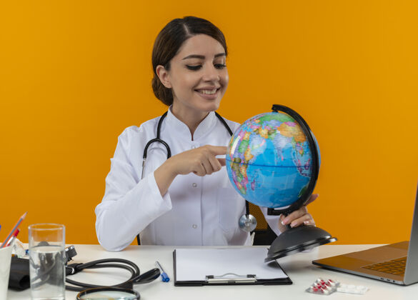 医生面带微笑的年轻女医生穿着医用长袍和听诊器坐在办公桌旁 拿着医疗工具和笔记本电脑 看着并用手指着隔离在黄色墙上的地球仪工具年轻人笔记本电脑
